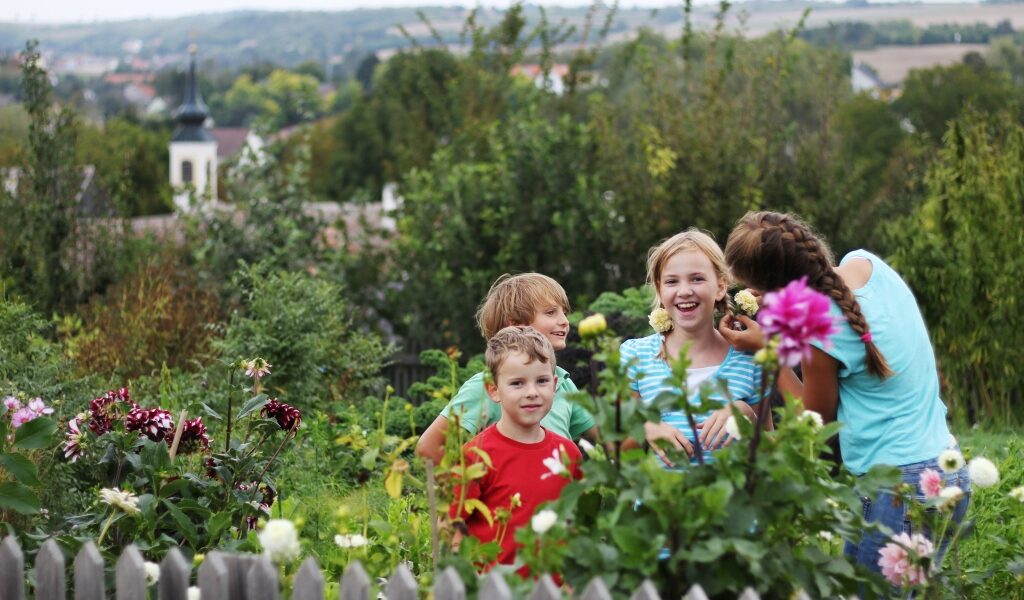 Vier Kinder stehen und lächeln zusammen in einem Garten mit blühenden Blumen und Grünzeug, mit einem Holzzaun im Vordergrund und einer ländlichen Landschaft im Hintergrund.