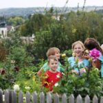 Vier Kinder stehen und lächeln zusammen in einem Garten mit blühenden Blumen und Grünzeug, mit einem Holzzaun im Vordergrund und einer ländlichen Landschaft im Hintergrund.