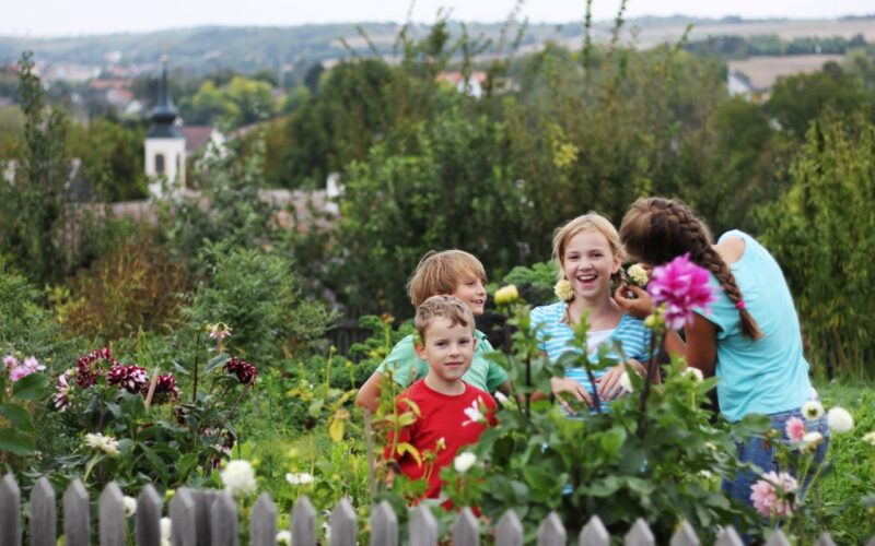 Vier Kinder stehen und lächeln zusammen in einem Garten mit blühenden Blumen und Grünzeug, mit einem Holzzaun im Vordergrund und einer ländlichen Landschaft im Hintergrund.