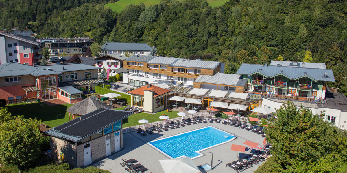 Luftaufnahme einer Hotelanlage mit Außenpool, Liegestühlen, Sonnenschirmen und grünen Hügeln und Bäumen in der Umgebung.