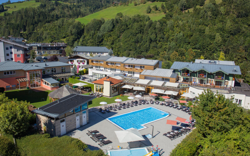 Luftaufnahme einer Hotelanlage mit Außenpool, Liegestühlen, Sonnenschirmen und grünen Hügeln und Bäumen in der Umgebung.