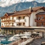 Ein modernes Alpenhotel mit Balkonen und einem Poolbereich, umgeben von Liegestühlen und Sonnenschirmen, vor dem Hintergrund bewaldeter Hügel und schroffer Berge.