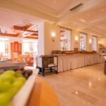 Ein geräumiger, gut beleuchteter Essbereich mit einem Buffet, weiß gedeckten Tischen und an den Wänden angeordneten Tabletts mit Speisen und Getränken.