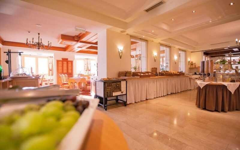 Ein geräumiger, gut beleuchteter Essbereich mit einem Buffet, weiß gedeckten Tischen und an den Wänden angeordneten Tabletts mit Speisen und Getränken.