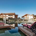 Ein luxuriöses Hotel mit Holzbalkonen und roten Liegestühlen umgibt einen spiegelnden Außenpool vor dem Hintergrund eines strahlend blauen Himmels und grüner Hügel.