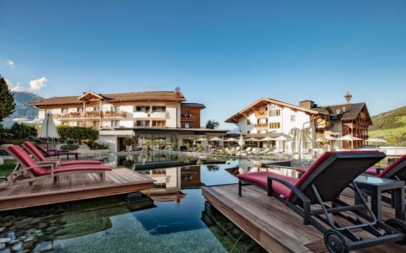 Ein luxuriöses Hotel mit Holzbalkonen und roten Liegestühlen umgibt einen spiegelnden Außenpool vor dem Hintergrund eines strahlend blauen Himmels und grüner Hügel.