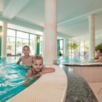 Zwei Kinder schwimmen in einem Innenpool, während zwei Erwachsene und ein Kind in einem angeschlossenen Whirlpool sitzen. Große Fenster geben den Blick auf die grüne Umgebung frei.