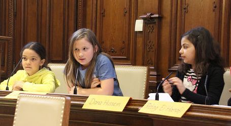 Drei Mädchen sitzen an einem hölzernen Konferenztisch mit Mikrofonen und nehmen an einer formellen Diskussion teil. Vor ihnen sind Namensschilder zu sehen.