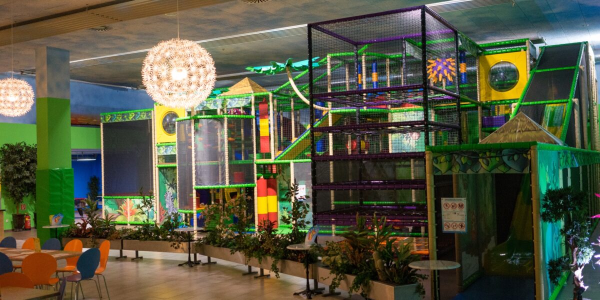 Indoor-Spielplatz mit farbenfrohen Klettergerüsten und Rutschen, umgeben von Pflanzen und einem Sitzbereich mit Tischen und Stühlen, unter hängenden dekorativen Lampen.