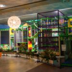 Indoor-Spielplatz mit farbenfrohen Klettergerüsten und Rutschen, umgeben von Pflanzen und einem Sitzbereich mit Tischen und Stühlen, unter hängenden dekorativen Lampen.