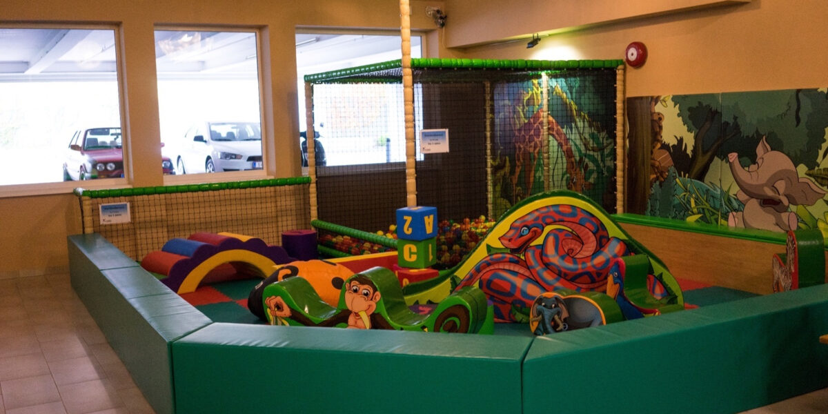 Indoor-Kinderspielbereich mit gepolsterten grünen Wänden, weichen Kletterstrukturen, bunten Schaumstoffblöcken und Dekorationen zum Thema Dschungel. Drei Fenster zeigen geparkte Autos im Freien.