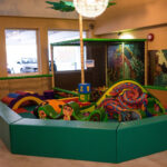 Indoor-Kinderspielbereich mit gepolsterten grünen Wänden, weichen Kletterstrukturen, bunten Schaumstoffblöcken und Dekorationen zum Thema Dschungel. Drei Fenster zeigen geparkte Autos im Freien.