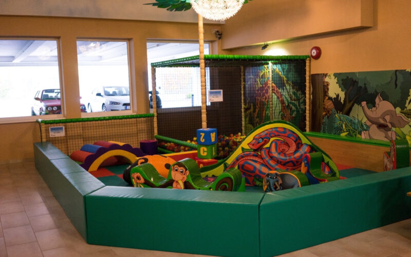 Indoor-Kinderspielbereich mit gepolsterten grünen Wänden, weichen Kletterstrukturen, bunten Schaumstoffblöcken und Dekorationen zum Thema Dschungel. Drei Fenster zeigen geparkte Autos im Freien.