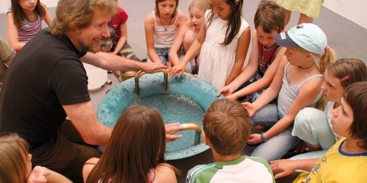 Ein Erwachsener und eine Gruppe von Kindern sitzen im Kreis um ein blaues Becken, legen ihre Hände auf den Rand und beobachten das Wasser darin.