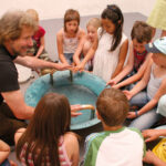 Ein Erwachsener und eine Gruppe von Kindern sitzen im Kreis um ein blaues Becken, legen ihre Hände auf den Rand und beobachten das Wasser darin.