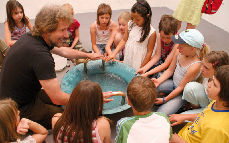 Ein Erwachsener und eine Gruppe von Kindern sitzen im Kreis um ein blaues Becken, legen ihre Hände auf den Rand und beobachten das Wasser darin.