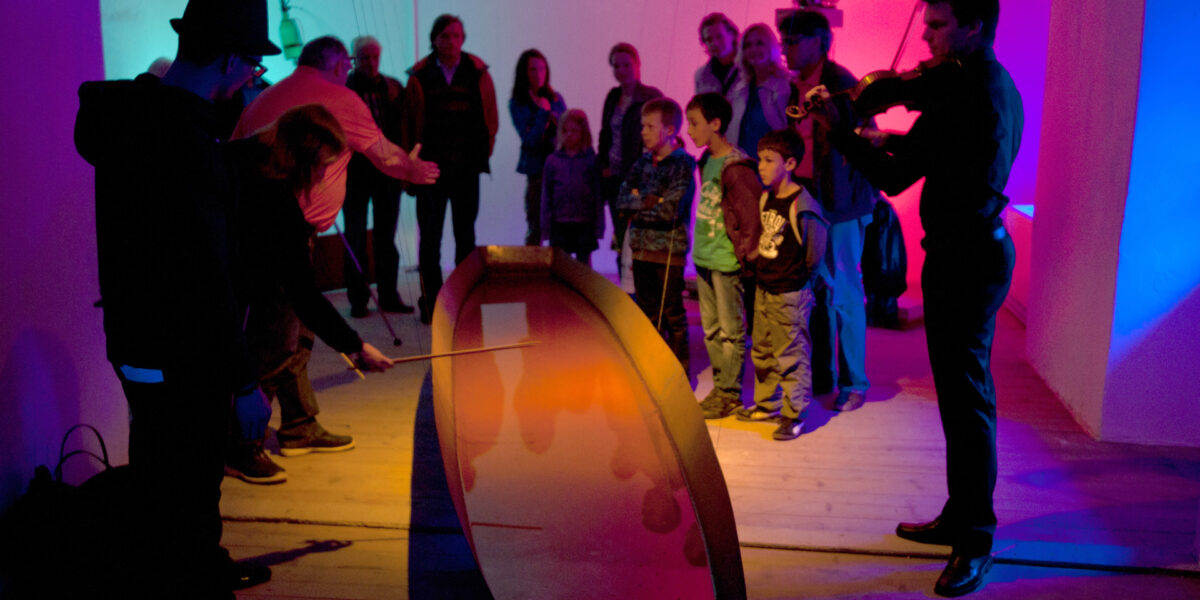 Eine Gruppe von Personen, darunter auch Kinder, betrachtet ein interaktives Exponat mit farbiger Beleuchtung, während ein Mann Geige spielt und eine andere Person das Exponat vorführt.