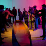 Eine Gruppe von Personen, darunter auch Kinder, betrachtet ein interaktives Exponat mit farbiger Beleuchtung, während ein Mann Geige spielt und eine andere Person das Exponat vorführt.