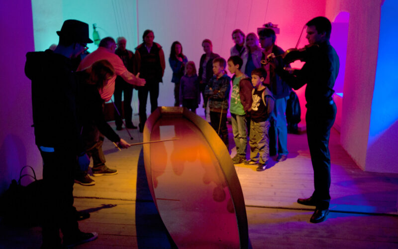 Eine Gruppe von Personen, darunter auch Kinder, betrachtet ein interaktives Exponat mit farbiger Beleuchtung, während ein Mann Geige spielt und eine andere Person das Exponat vorführt.