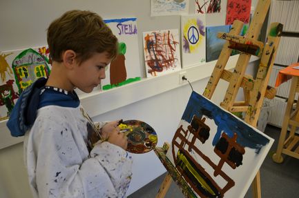 Ein kleiner Junge hält eine Farbpalette und einen Pinsel in der Hand und malt in einem Kunstunterrichtsraum ein Bild von einem Zaun und Bäumen auf eine Leinwand. An der Wand hinter ihm hängen weitere Gemälde.