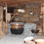 Eine rustikale Küche mit Holzwänden, einem großen schwarzen Kessel aus Stein, verschiedenen Kochgeräten aus Holz und Metall und Fässern.