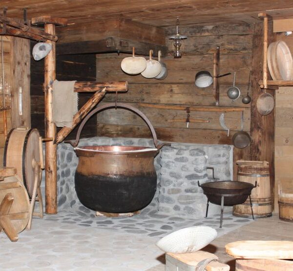 Eine rustikale Küche mit Holzwänden, einem großen schwarzen Kessel aus Stein, verschiedenen Kochgeräten aus Holz und Metall und Fässern.