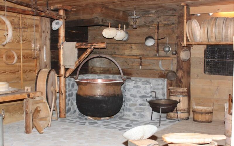 Eine rustikale Küche mit Holzwänden, einem großen schwarzen Kessel aus Stein, verschiedenen Kochgeräten aus Holz und Metall und Fässern.