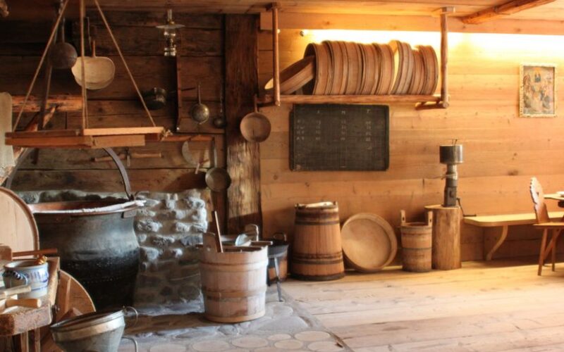 Ein rustikaler Holzraum mit Fässern, Wannen, hängenden Töpfen und Küchengeräten, mit Steinmetzarbeiten um einen großen Kessel und Regalen mit runden Holztabletts.