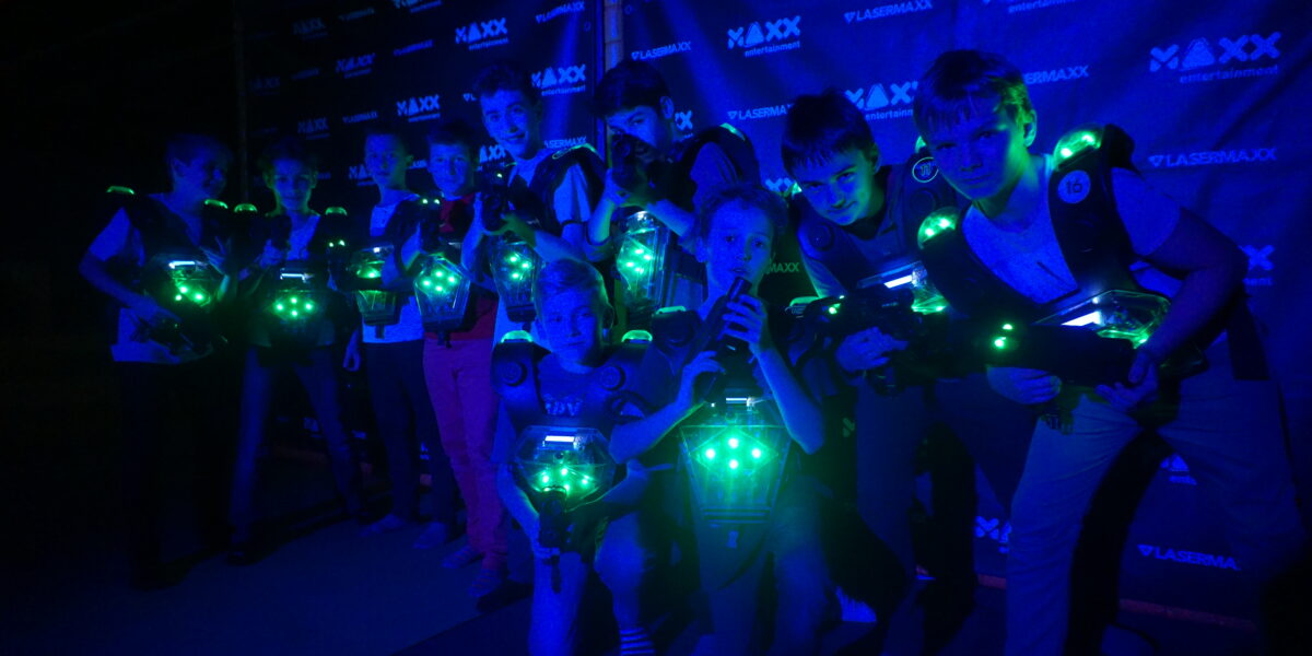 Eine Gruppe von Personen, die Laser-Tag-Westen tragen und Laser-Tag-Pistolen in der Hand halten, posiert unter blauem Licht vor einem Markenhintergrund.