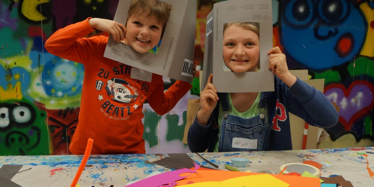 Zwei Kinder sitzen an einem mit buntem Papier und Bastelmaterial bedeckten Tisch, halten sich Papprahmen vors Gesicht und lächeln in die Kamera. An der Wand hinter ihnen ist ein Graffiti-Wandbild zu sehen.