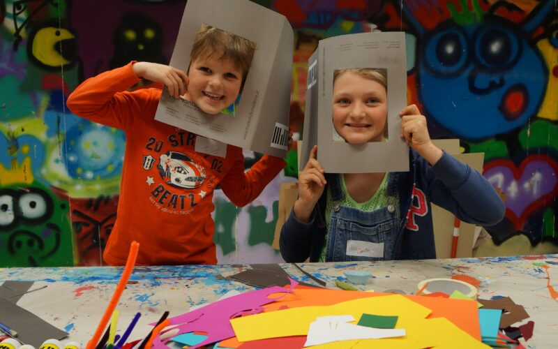 Zwei Kinder sitzen an einem mit buntem Papier und Bastelmaterial bedeckten Tisch, halten sich Papprahmen vors Gesicht und lächeln in die Kamera. An der Wand hinter ihnen ist ein Graffiti-Wandbild zu sehen.