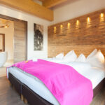 Ein modernes Hotelzimmer mit einem Doppelbett, weißer Bettwäsche, einer hellrosa Decke, einer Akzentwand aus Holz und einer offenen Tür, die in ein Badezimmer mit einer freistehenden Badewanne führt.