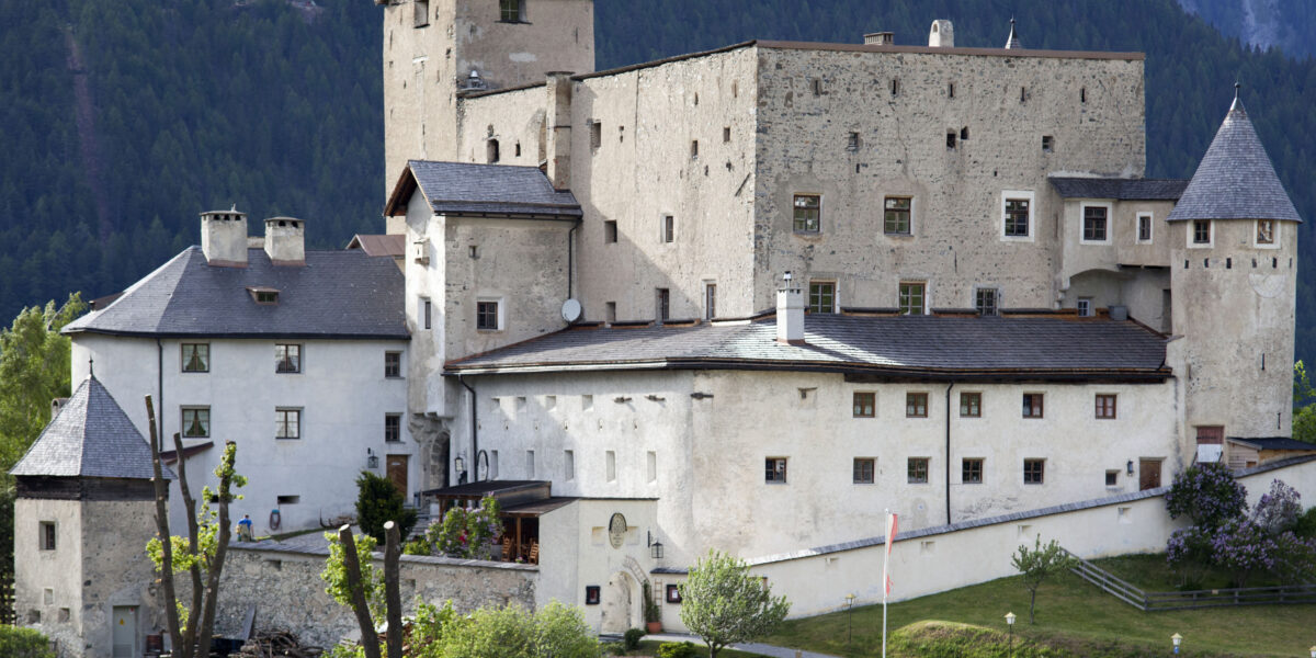 Eine große Steinburg mit mehreren Türmen und Schrägdächern liegt an einem bewaldeten Hang, vor dem ein Auto auf der Wiese geparkt ist.