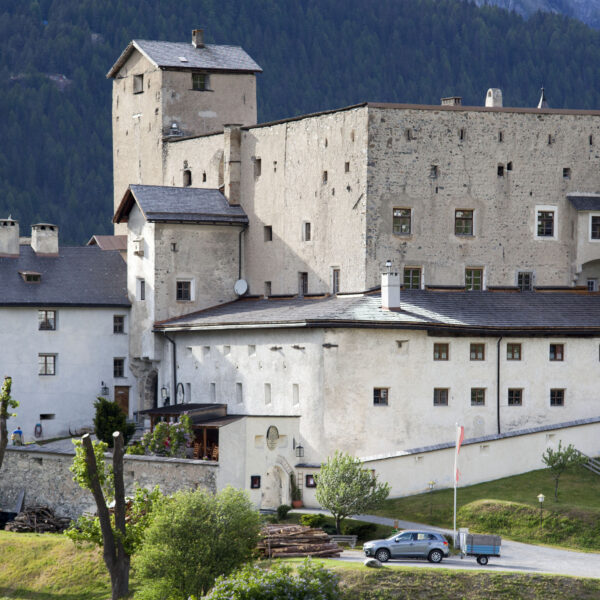 Eine große Steinburg mit mehreren Türmen und Schrägdächern liegt an einem bewaldeten Hang, vor dem ein Auto auf der Wiese geparkt ist.