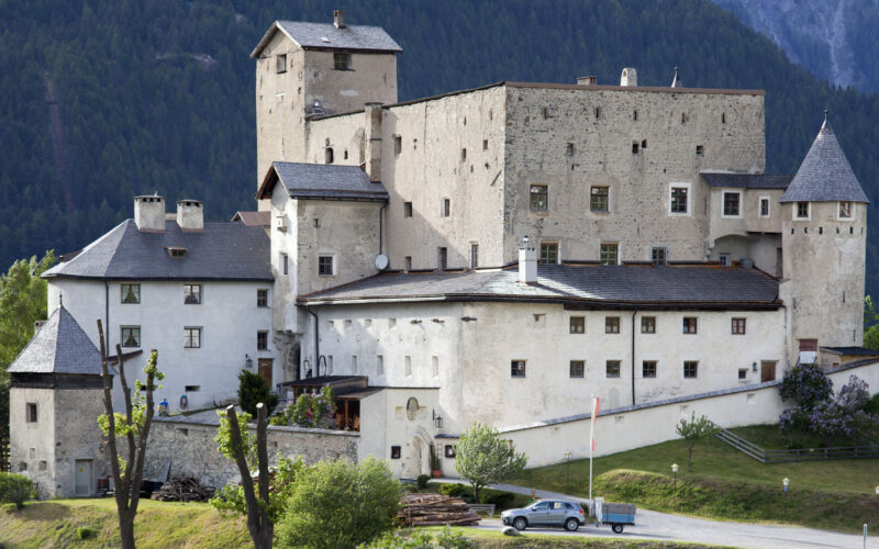 Eine große Steinburg mit mehreren Türmen und Schrägdächern liegt an einem bewaldeten Hang, vor dem ein Auto auf der Wiese geparkt ist.
