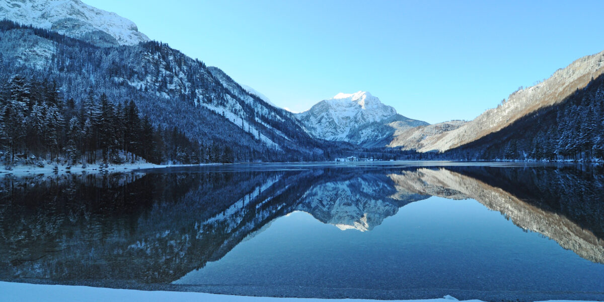 In einem ruhigen See spiegeln sich schneebedeckte Berge und Bäume unter einem klaren blauen Himmel wider, im Vordergrund ist das Ufer mit Schnee bedeckt.