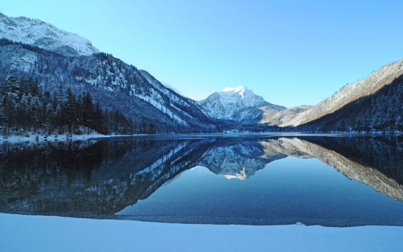 In einem ruhigen See spiegeln sich schneebedeckte Berge und Bäume unter einem klaren blauen Himmel wider, im Vordergrund ist das Ufer mit Schnee bedeckt.