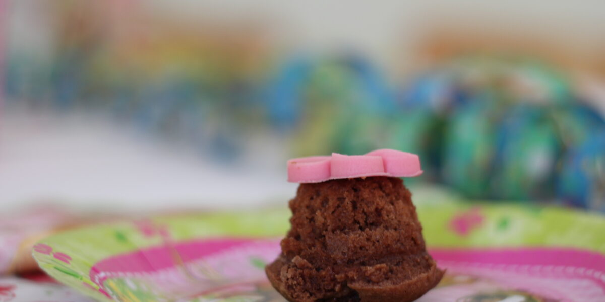 Ein kleiner Schokoladenkuchen mit rosa Fondantdekoration liegt auf einem bunten Pappteller mit unscharfem Hintergrund.