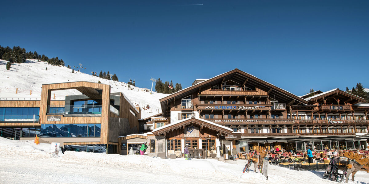 Die hölzernen Gebäude des Skigebiets mit Sitzgelegenheiten im Freien befinden sich am Fuße eines verschneiten Berghangs unter einem strahlend blauen Himmel.