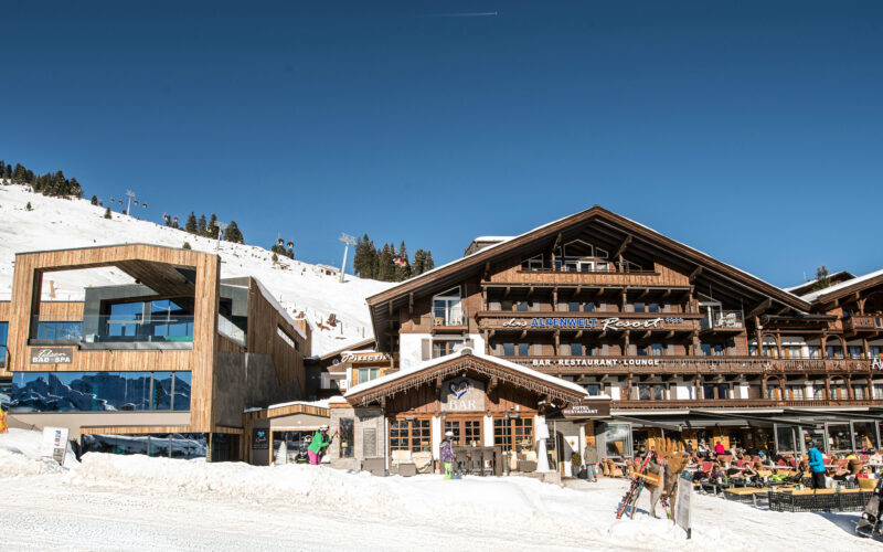 Die hölzernen Gebäude des Skigebiets mit Sitzgelegenheiten im Freien befinden sich am Fuße eines verschneiten Berghangs unter einem strahlend blauen Himmel.