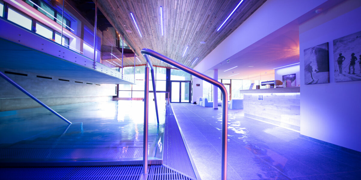 Moderner Hallenbadbereich mit blauer Beleuchtung, Holzdecke, Metallgeländern und einem Empfangstresen im Hintergrund.