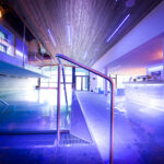 Moderner Hallenbadbereich mit blauer Beleuchtung, Holzdecke, Metallgeländern und einem Empfangstresen im Hintergrund.