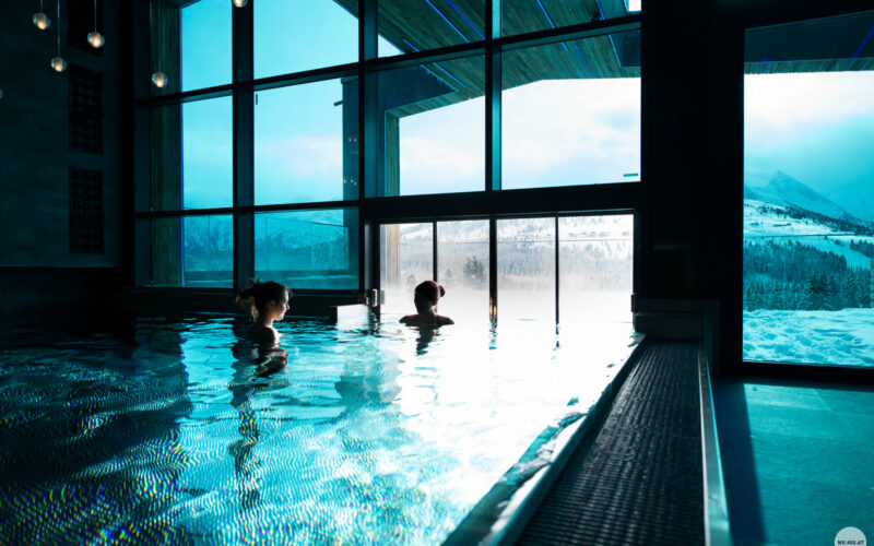 Zwei Personen entspannen sich in einem Hallenbad neben großen Fenstern, während draußen eine verschneite Berglandschaft zu sehen ist.