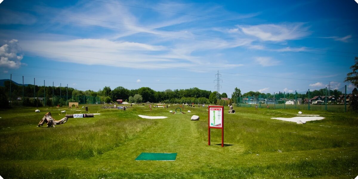 Ein grasbewachsener Discgolfplatz unter blauem Himmel mit vereinzelten Wolken, mit einem Abschlagplatz, Schildern, Sandfängen und Menschen in der Ferne.