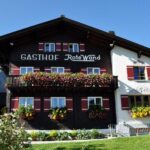 Ein traditionelles Alpengasthaus mit Blumenkästen unter den Fenstern und dem Schriftzug "Gasthof Rote Wand" an der Fassade, vor einem strahlend blauen Himmel.