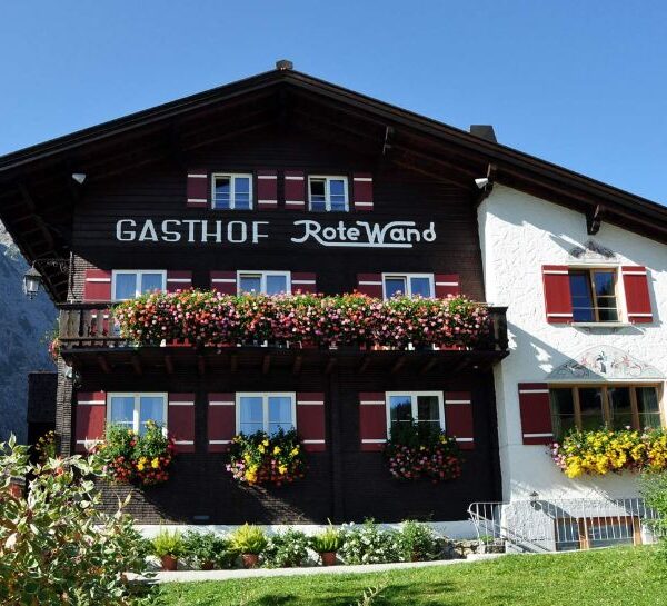 Ein traditionelles Alpengasthaus mit Blumenkästen unter den Fenstern und dem Schriftzug "Gasthof Rote Wand" an der Fassade, vor einem strahlend blauen Himmel.