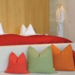Ein modernes Hotelzimmer mit einem Doppelbett, weißen Kissen, einer roten Decke, einem grauen Sofa mit bunten Kissen und einem weißen Bademantel, der an der Wand hängt.