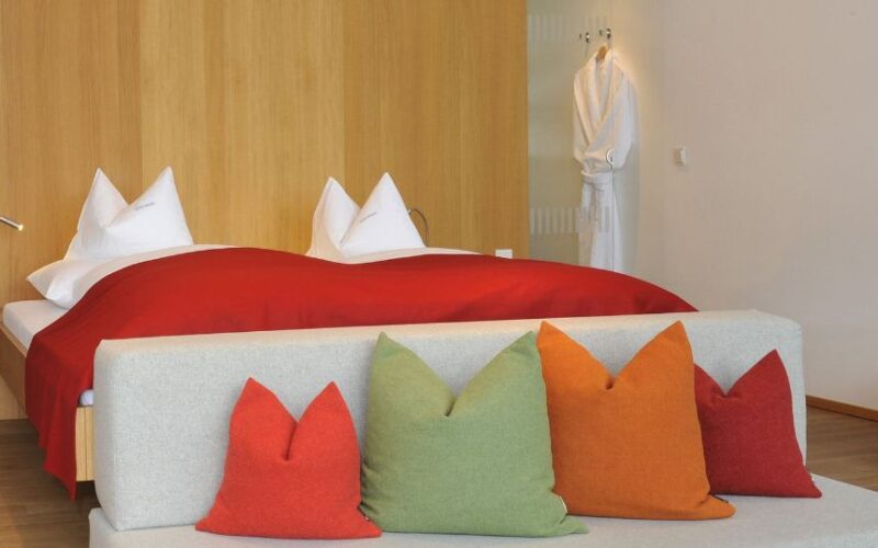 Ein modernes Hotelzimmer mit einem Doppelbett, weißen Kissen, einer roten Decke, einem grauen Sofa mit bunten Kissen und einem weißen Bademantel, der an der Wand hängt.