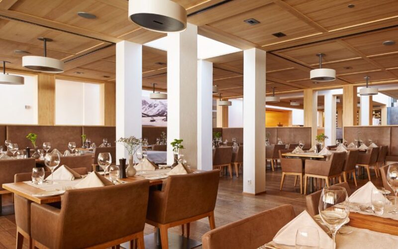 Moderner Essbereich in einem Restaurant mit braunen Lederstühlen, Holztischen mit Gläsern und Servietten, Holzdecke und weißen Säulen. Der Raum ist menschenleer.