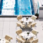 Außenpoolbereich mit runden Tischen und Stühlen, die mit weißem Fell bezogen sind und auf einer gefliesten Terrasse neben einem rechteckigen Swimmingpool stehen.